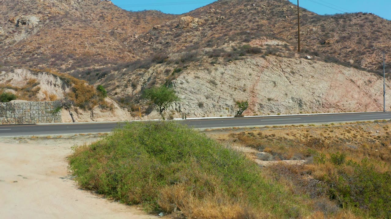 The road is empty at Marquis Los Cabos, Mexico, with foreground desert scrub, sandy ground, a stone wall, utility poles, rocky hills, and no visible people.
