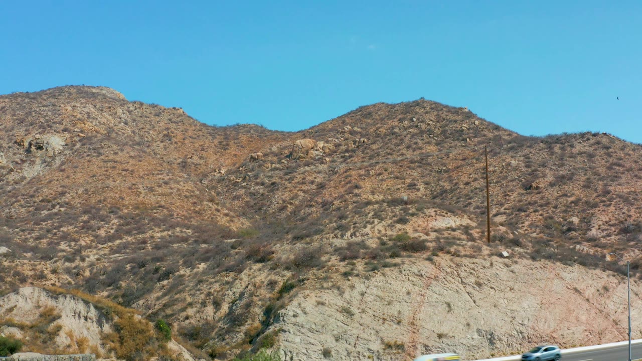 A white car moves along the Marquis Los Cabos highway while dry rocky hills, utility poles, and a clear blue sky fill the background, with no visible people.