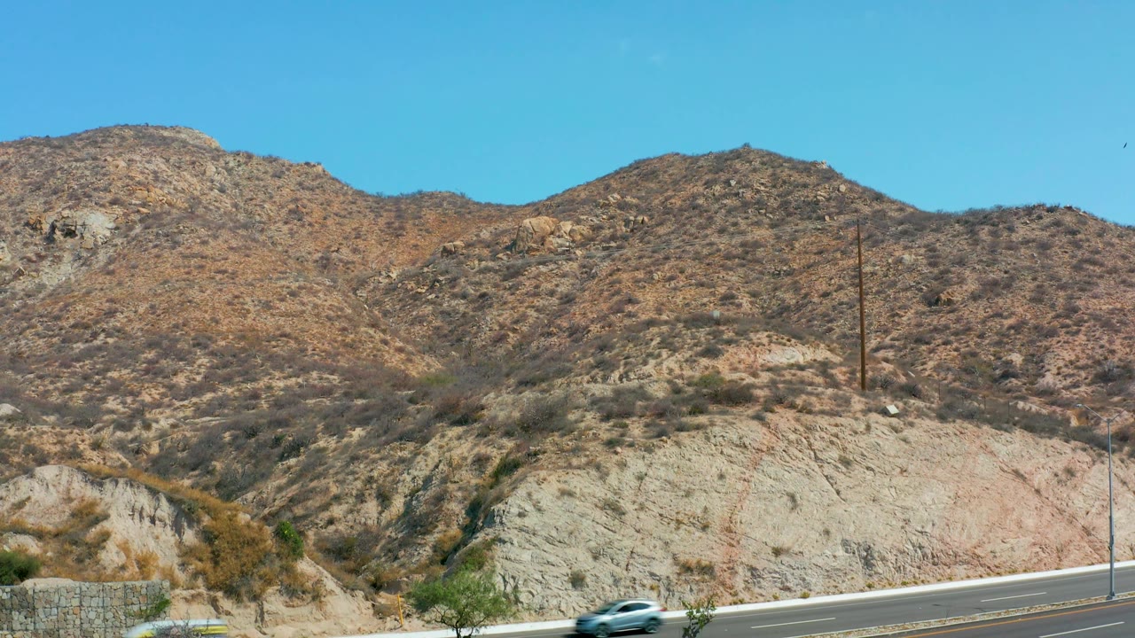 A light-colored car travels across the lower road in Marquis Los Cabos, backed by layered tan hills, scrub vegetation, and blue sky, with no visible people.