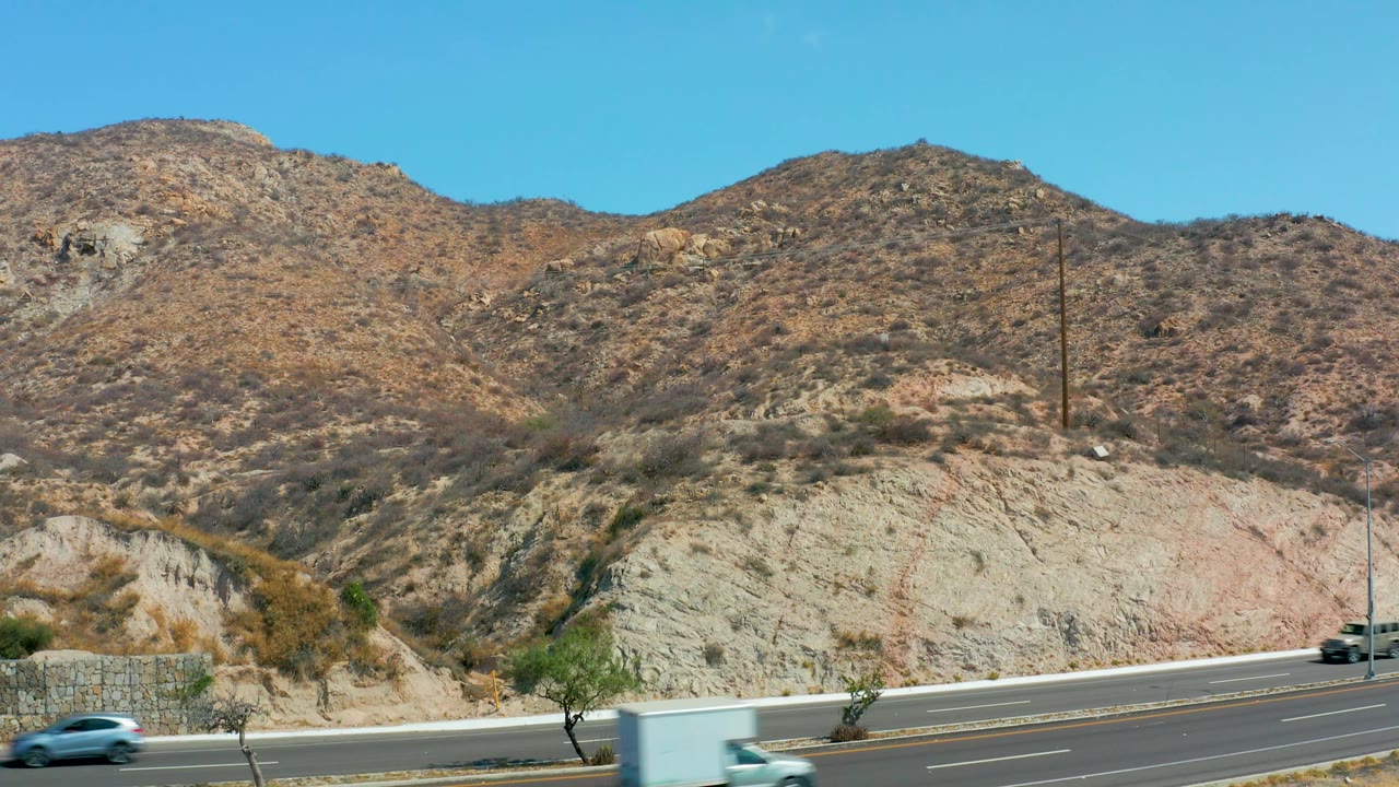 A white box truck and a small car pass along the Marquis Los Cabos highway beneath dry hills and roadside trees, with no visible people in the frame.