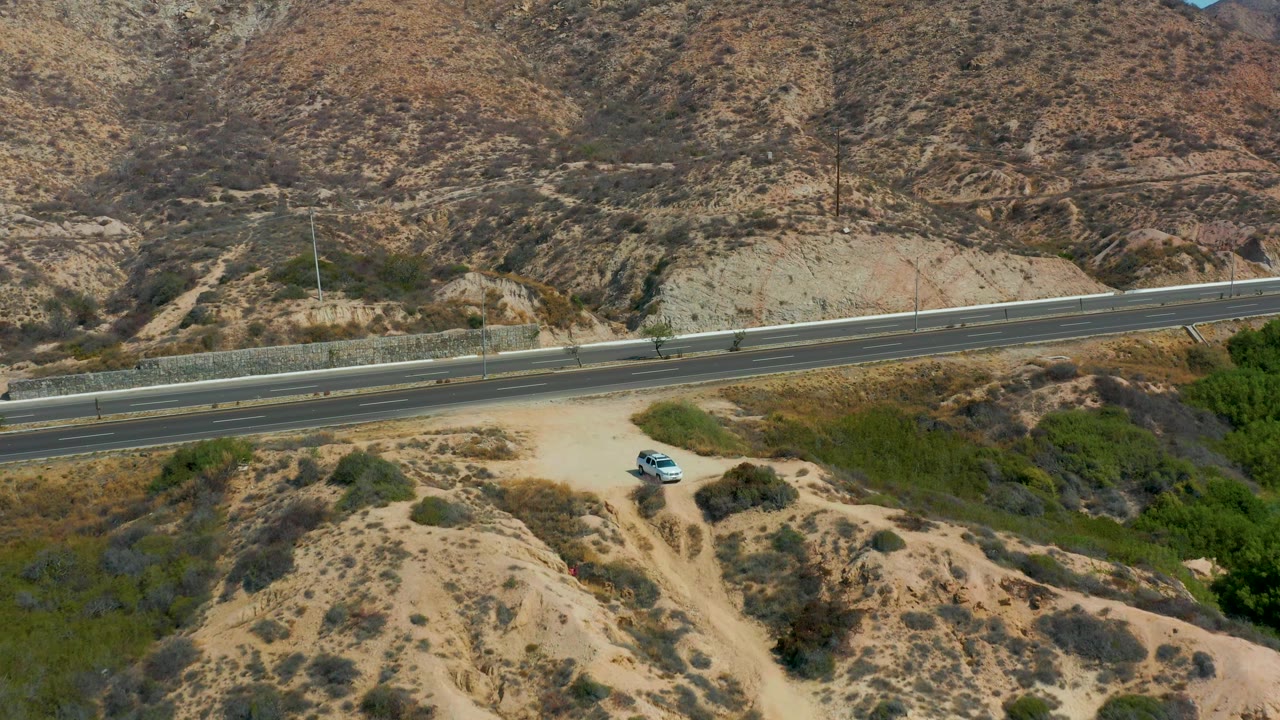 The second frame keeps the parked white car low in the shot while the highway spans the middle ground below rocky desert hills near Marquis Los Cabos, with no visible people.