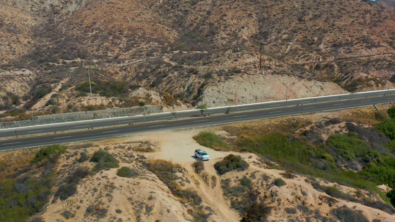 Aerial footage near Marquis Los Cabos shows the highway cutting across dry terrain, the parked white car set back on the dirt path, scattered shrubs, steep rocky hills, and no visible people.