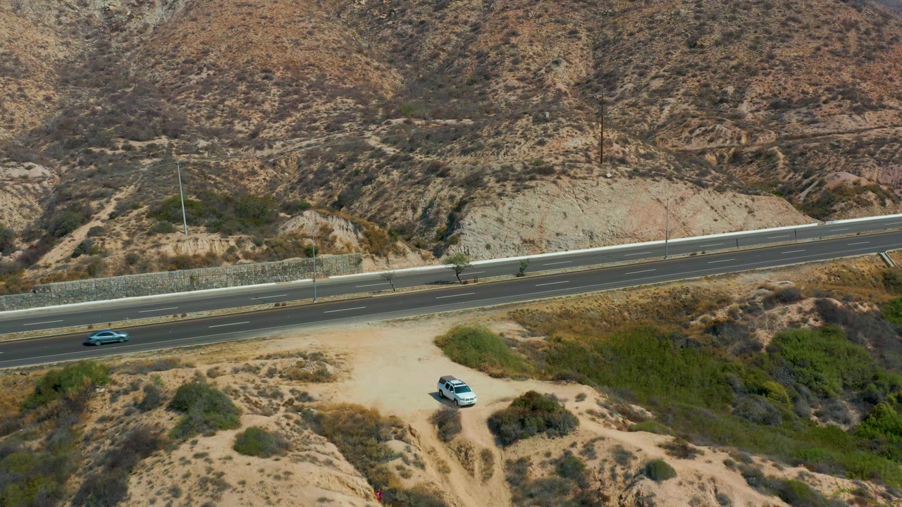A blue vehicle enters the highway scene above the parked white car on the sandy track, with dry hillsides and sparse vegetation around Marquis Los Cabos and no visible people.