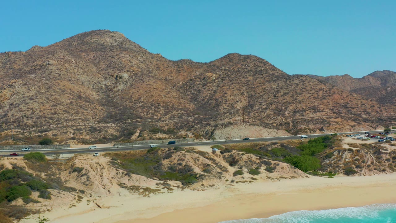 Wide elevated view at Marquis Los Cabos, Mexico showing sandy beach, turquoise wave edge, coastal highway traffic, arid mountains, clear blue sky, and no visible people.
