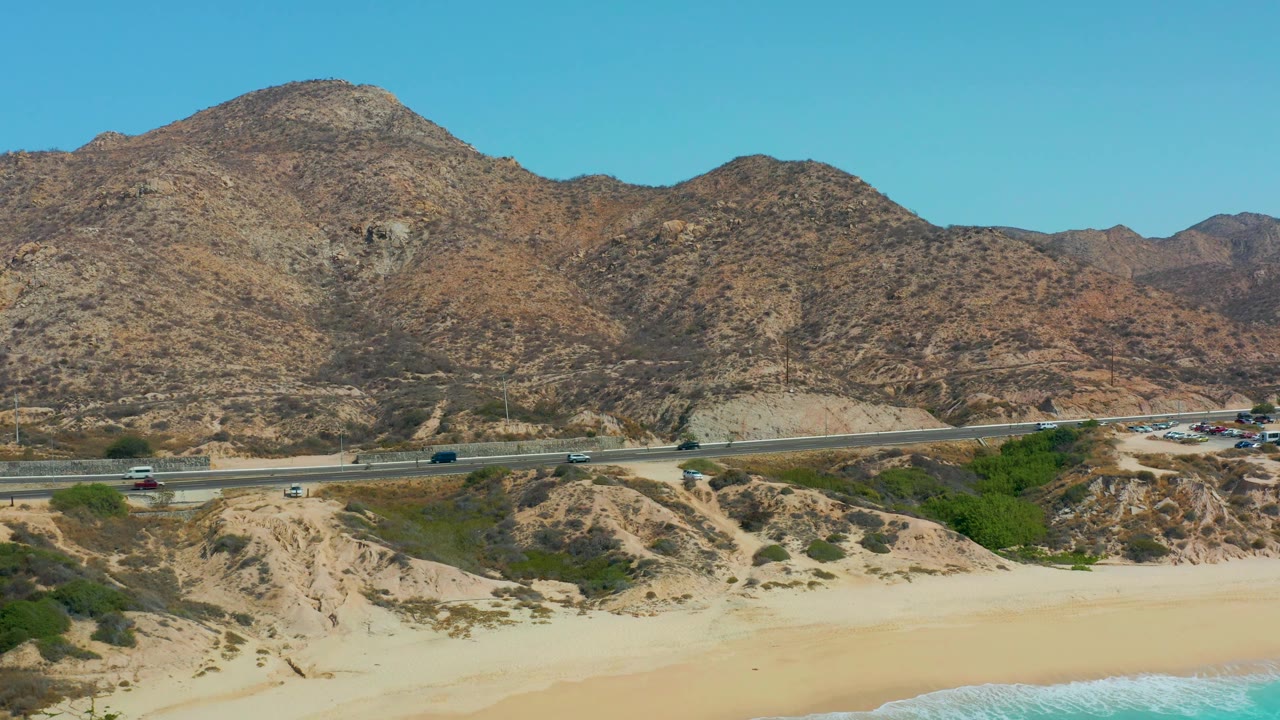At 0:02, the wide aerial view shows the coastal highway crossing above the beach, with small vehicles on the lanes, rocky brown hills behind, and no visible people.