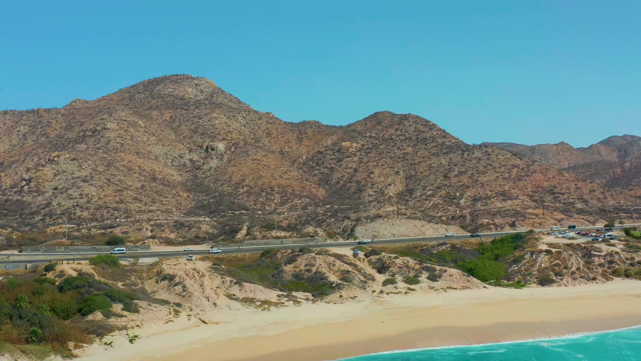 Opening frame of Marquis Los Cabos, Mexico showing turquoise water, sandy beach, a coastal highway with small vehicles, rocky mountains, clear blue sky, and no visible people.