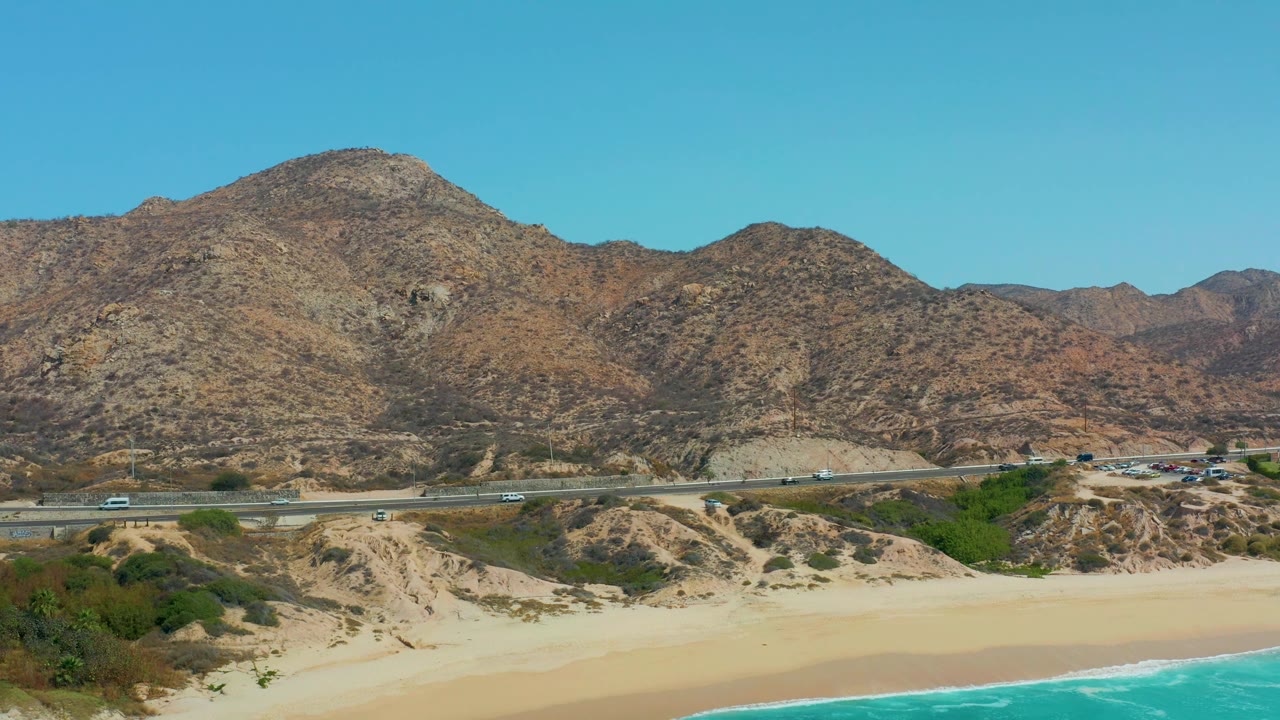 At 0:02, the frame shows the same Marquis Los Cabos coast with the turquoise ocean cropped along the bottom, a few vehicles on the highway, mountain slopes behind, and no visible people.