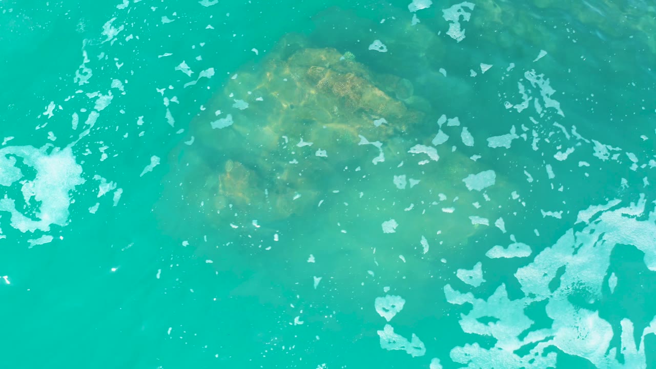 At Marquis Los Cabos, the submerged rock remains visible under clear turquoise water as scattered foam patches spread across the lower half, with no visible people.
