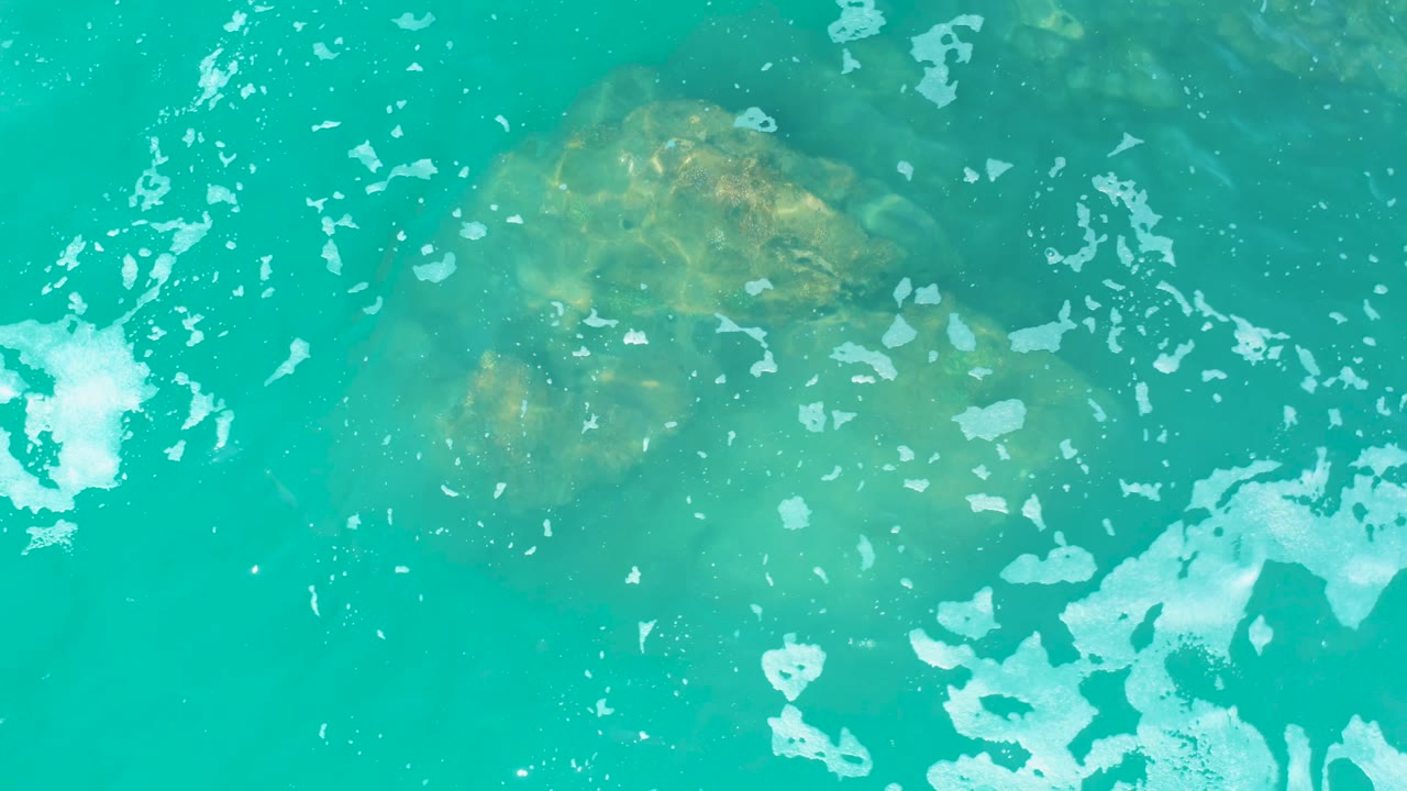 At Marquis Los Cabos, an overhead view shows the rock softened by water glare near the top and a larger white foam patch entering from the right, with no visible people.