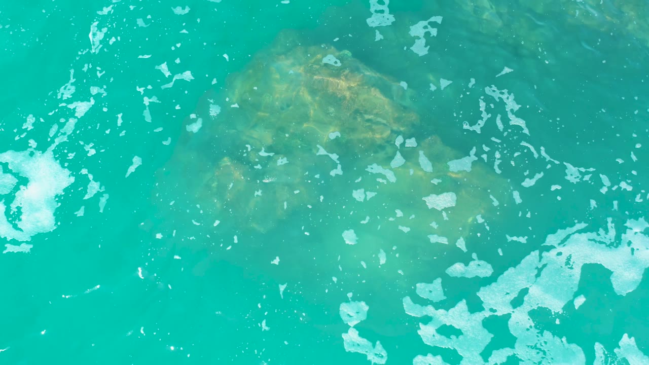 At Marquis Los Cabos, the rock sits beneath green-blue water near the upper left while white foam appears along the left and lower right edges, with no visible people.