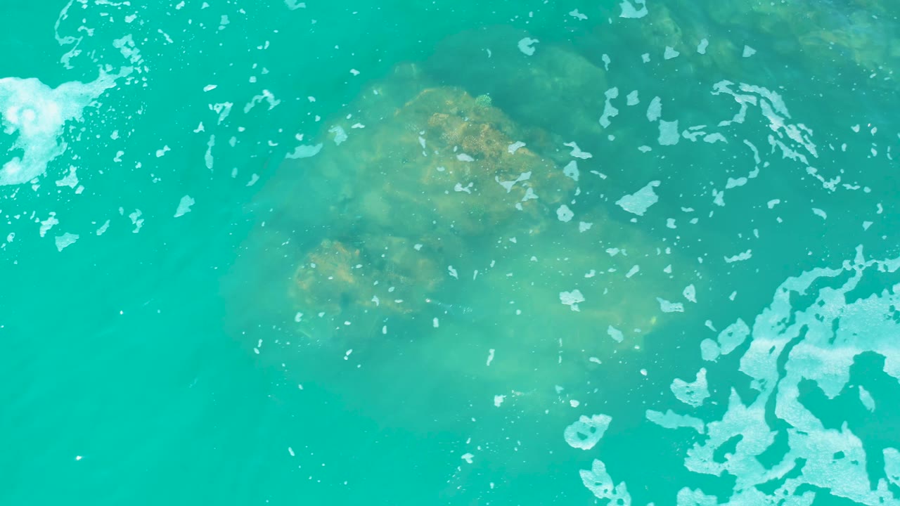 At Marquis Los Cabos, Mexico, the submerged rock sits under clear turquoise water while a larger patch of white foam gathers toward the right side, with no visible people.