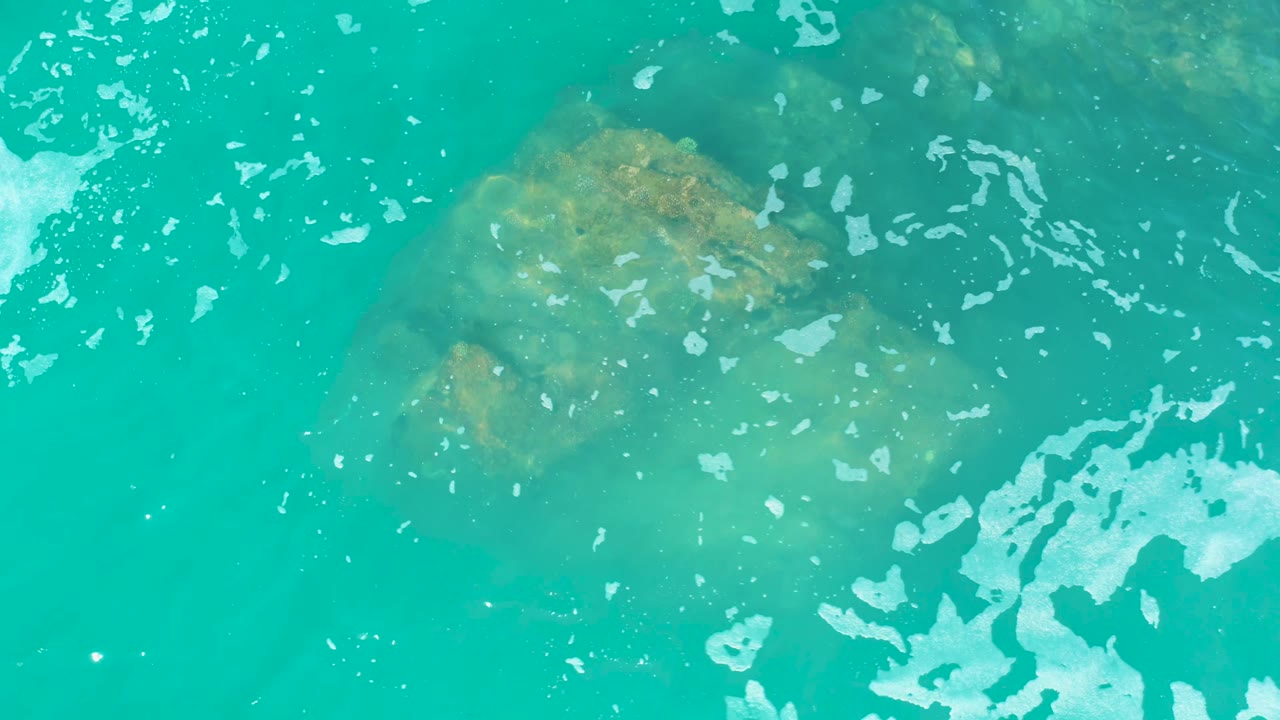The Marquis Los Cabos frame shows the rock larger in view beneath turquoise water, with white foam along the lower right and no visible people.
