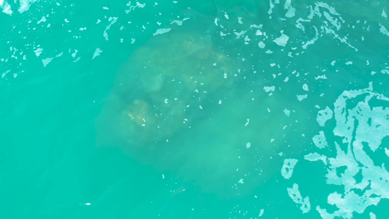 At Marquis Los Cabos, Mexico, the turtle is barely visible beneath blue-green water, with a broad white foam patch at the right edge and no visible people.