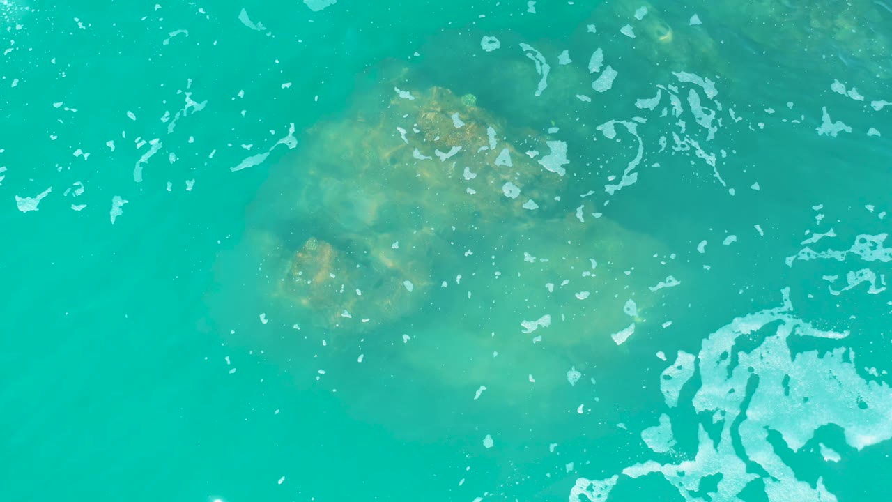 Turquoise ocean surface with jellyfish below, bubbles, and a pale foam patch at lower right.
