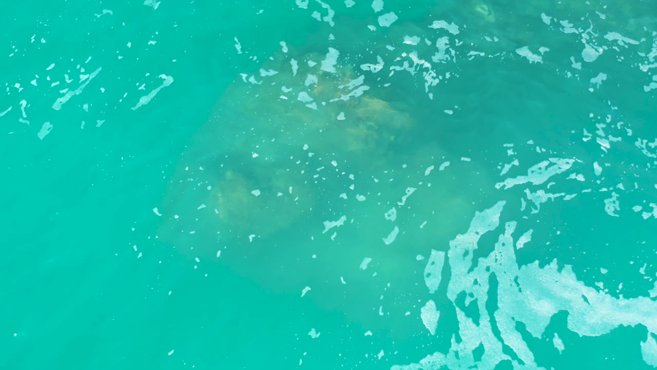 A muted manatee outline is visible under the turquoise sea surface at Marquis Los Cabos, Mexico, with broken foam flecks across the top and right side and no visible people.
