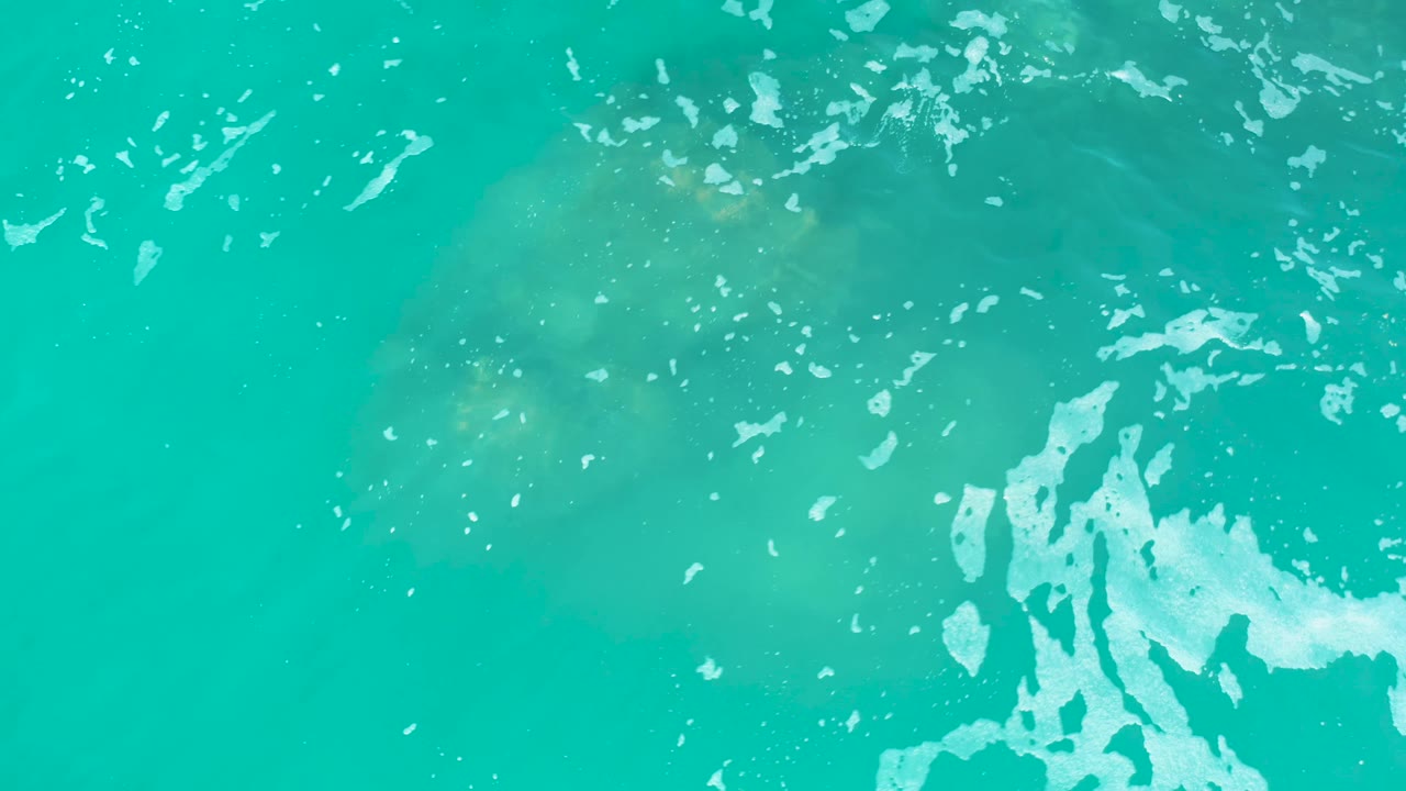 The manatee appears faint beneath rippled turquoise water at Marquis Los Cabos, Mexico, with white foam crossing the upper area and no visible people.