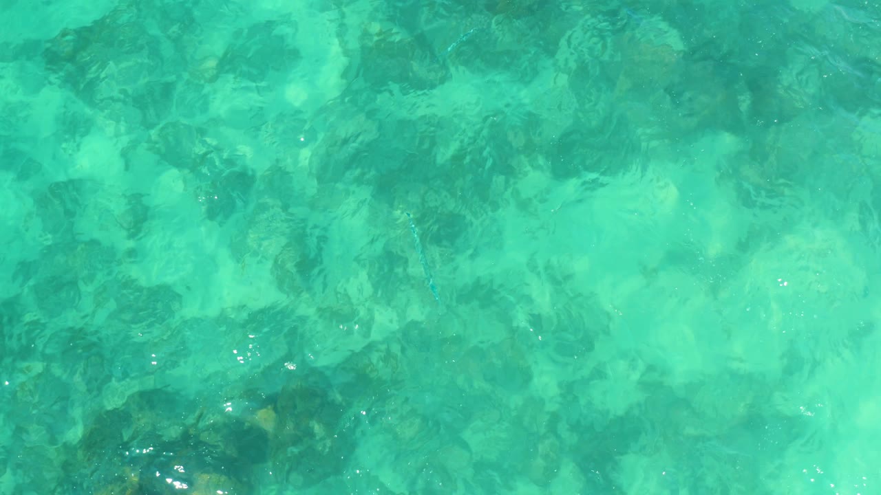 Aqua water at Marquis Los Cabos shows darker underwater rock clusters softened by small surface ripples, with no visible people.