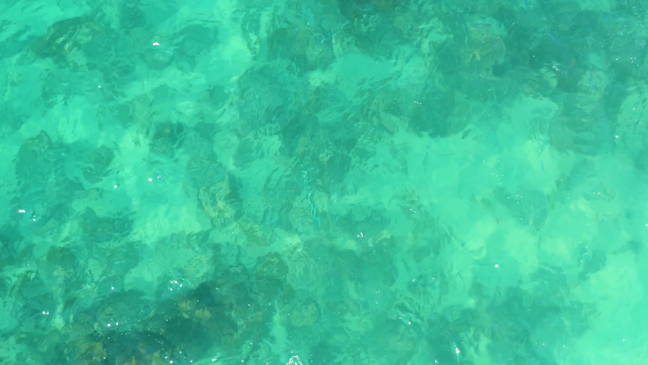 Green-blue water at Marquis Los Cabos passes over mottled submerged rocks, with brighter ripples crossing the frame and no visible people.