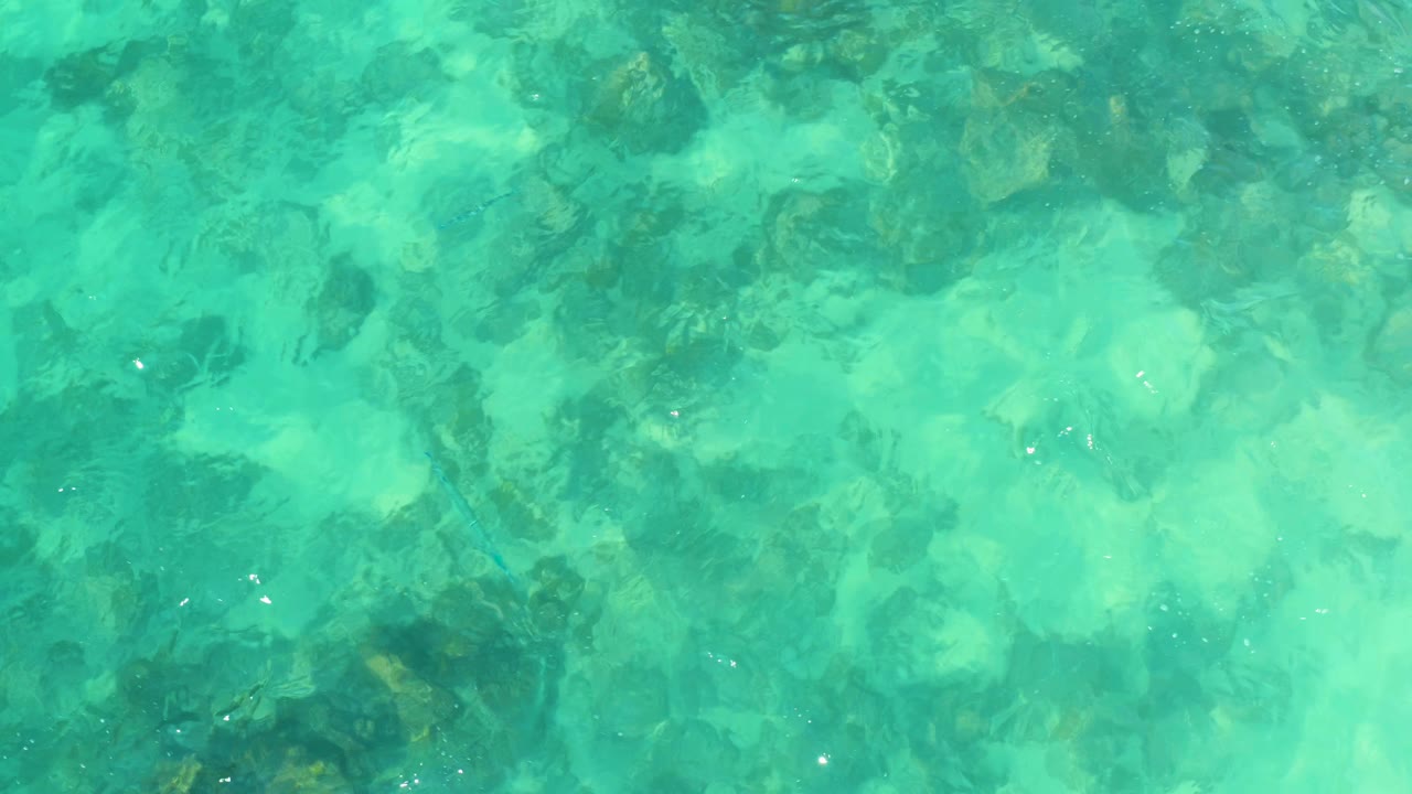 Opening frame of clear turquoise water at Marquis Los Cabos, Mexico, with dark rocky patches visible below the rippled surface and no visible people.