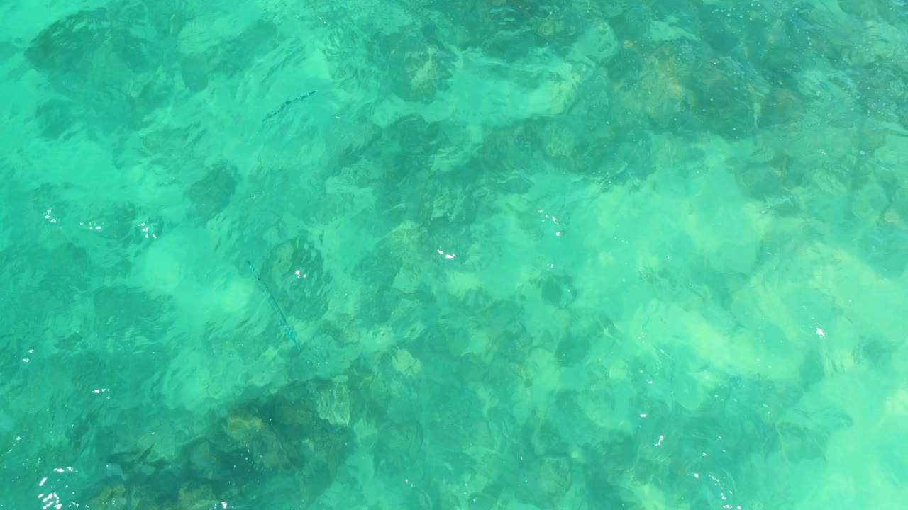 Turquoise ocean water at Marquis Los Cabos, Mexico, ripples across a mottled rocky seabed with scattered bright reflections and no visible people.