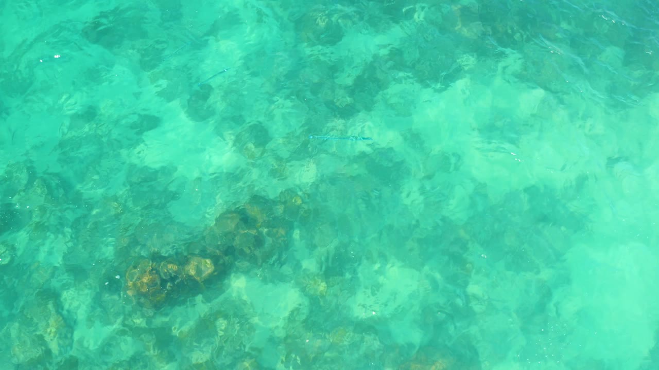Shallow turquoise water at Marquis Los Cabos, Mexico, with mottled rocks and pale seabed areas visible below the rippled surface and no visible people.