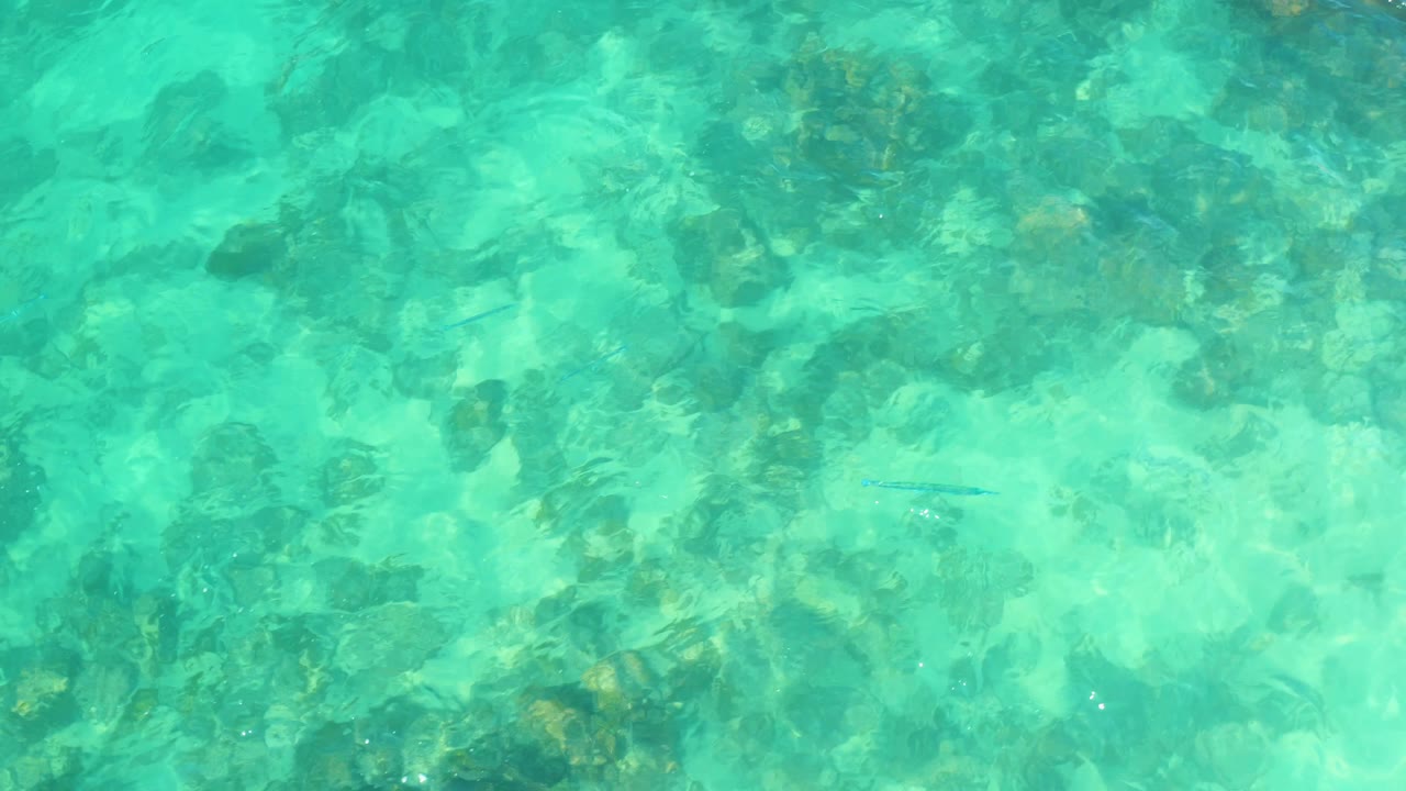 Shallow turquoise ocean water reveals darker submerged rock clusters and lighter sandy areas at Marquis Los Cabos, Mexico, with no visible people.