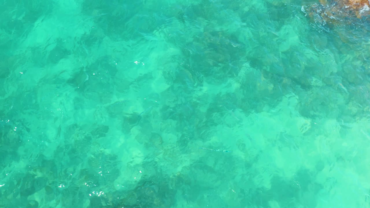Overhead frame from Marquis Los Cabos, Mexico, showing shallow aqua water, darker rock patches below, and small bright surface glints with no visible people.