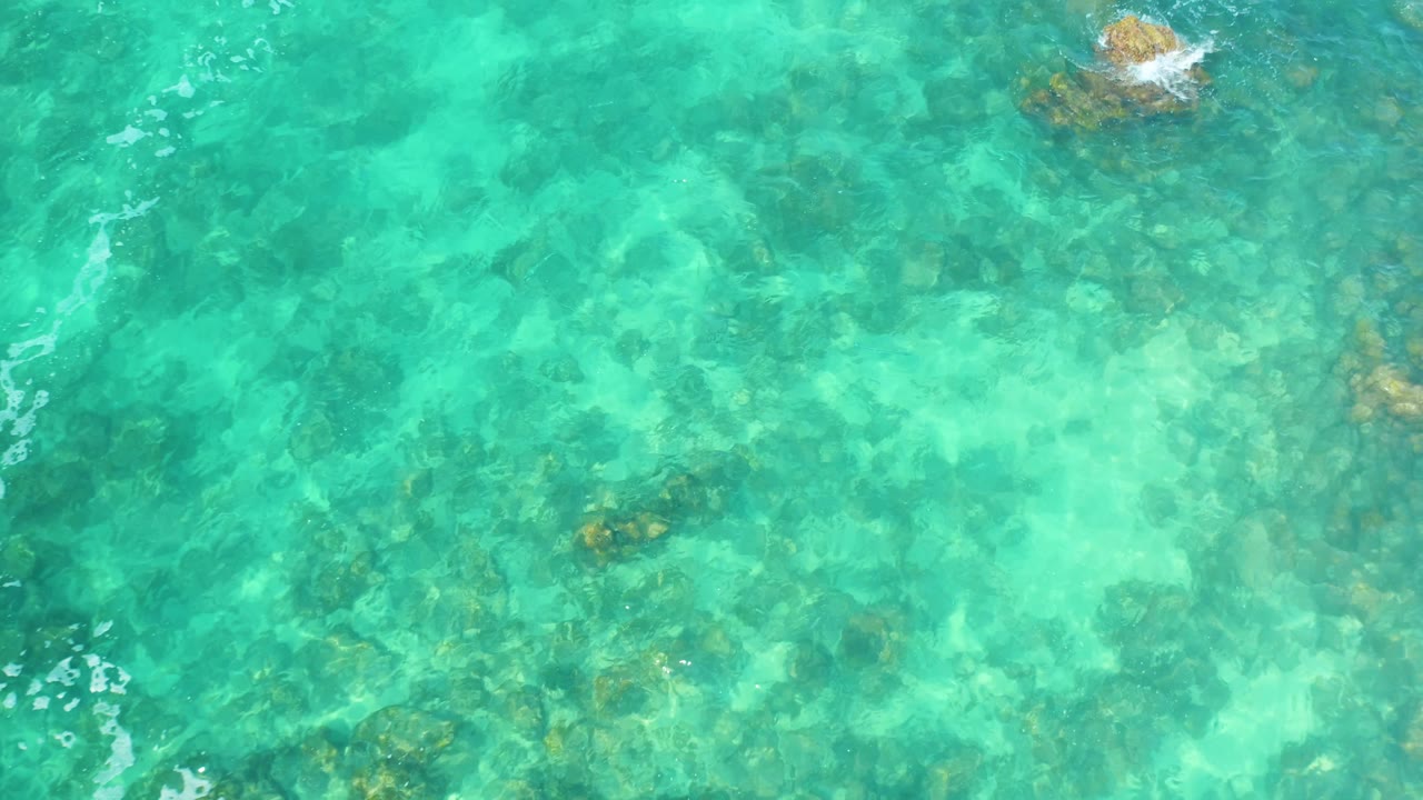 An overhead frame at Marquis Los Cabos, Mexico, shows clear water and a small rock near the top edge creating white foam, with no visible people.