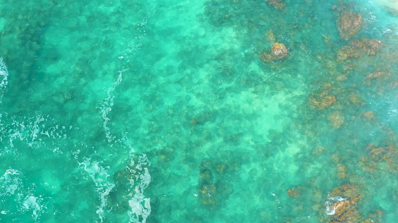 Overhead view of clear turquoise water at Marquis Los Cabos with submerged rocks near the upper right and a thin foam line along the left edge, no visible people.