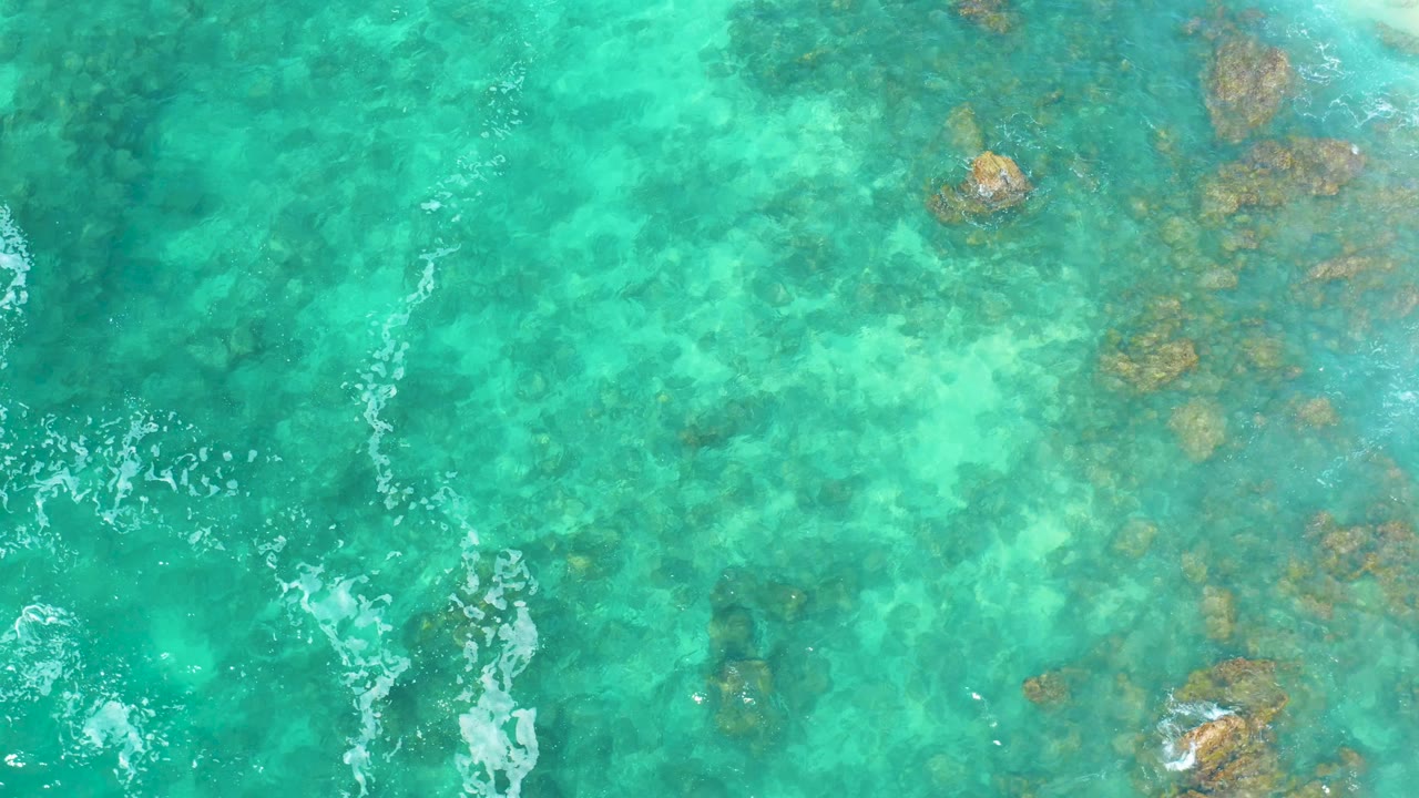 Overhead Marquis Los Cabos water view with pale turquoise shallows, scattered underwater rocks, and a narrow white foam streak entering from the lower left, no visible people.