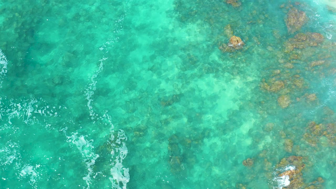 Top-down frame of turquoise ocean at Marquis Los Cabos with a cluster of brown rocks on the right and broken white foam crossing the left side, no visible people.