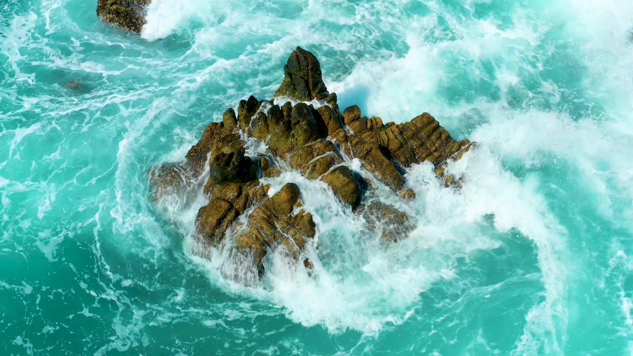 Turquoise waves breaking around a jagged rocky outcrop.