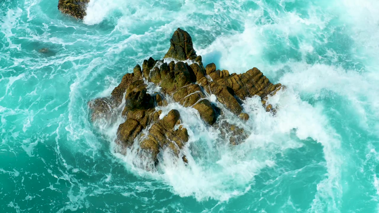 Aerial view of white surf curling around the brown rock formation in turquoise water at Marquis Los Cabos, Mexico, with no visible people.