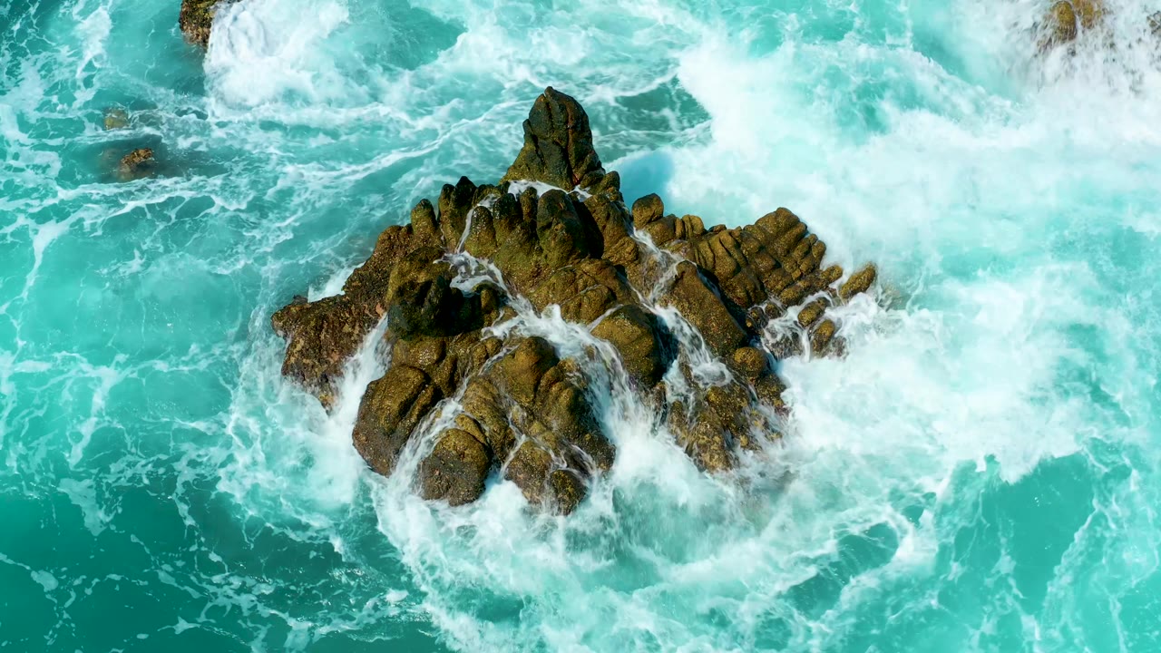 Foam spreads across the right side of the central rocky outcrop while turquoise water surrounds the formation at Marquis Los Cabos, Mexico, with no visible people.