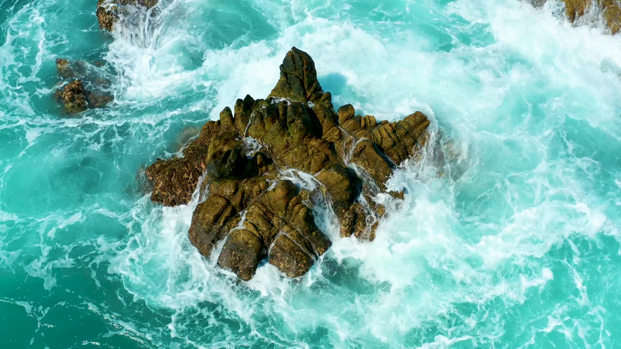 An overhead frame shows surf washing over the upper and right edges of dark brown rocks in turquoise water at Marquis Los Cabos, Mexico, with no visible people.