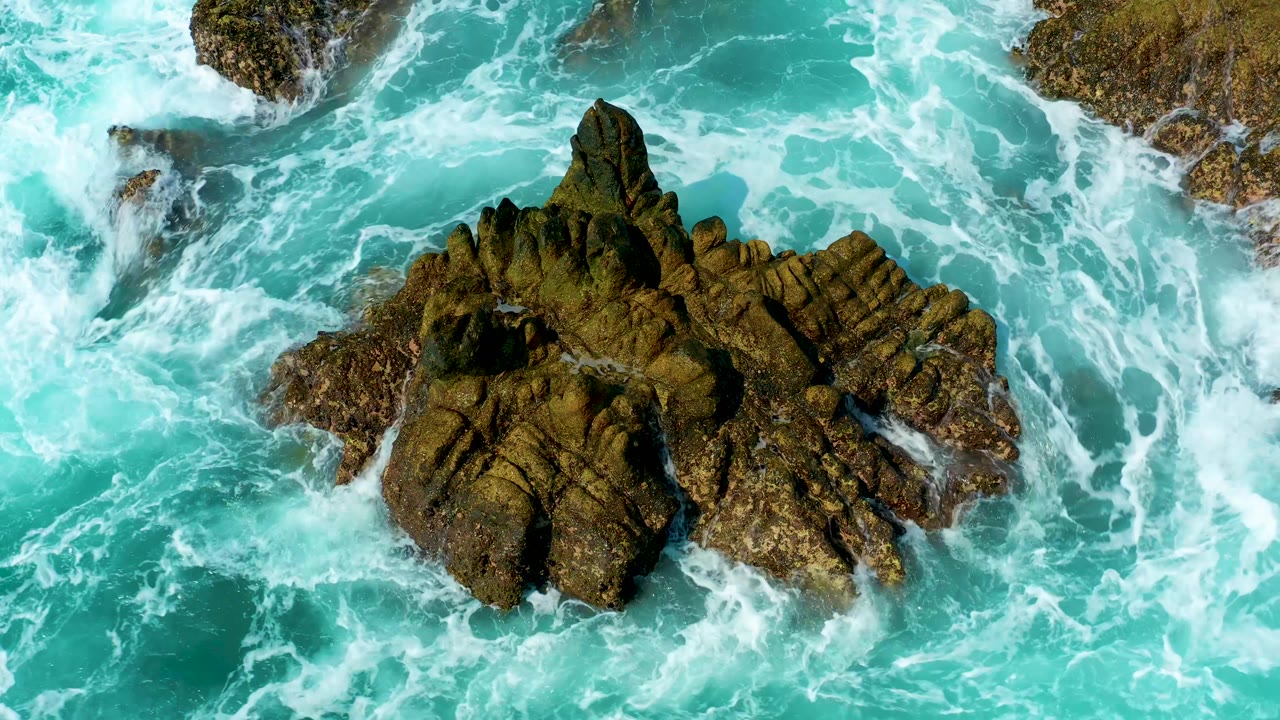 Aerial frame from Marquis Los Cabos, Mexico, with white water curling around the central dark rock while turquoise water fills the surrounding frame, with no visible people.