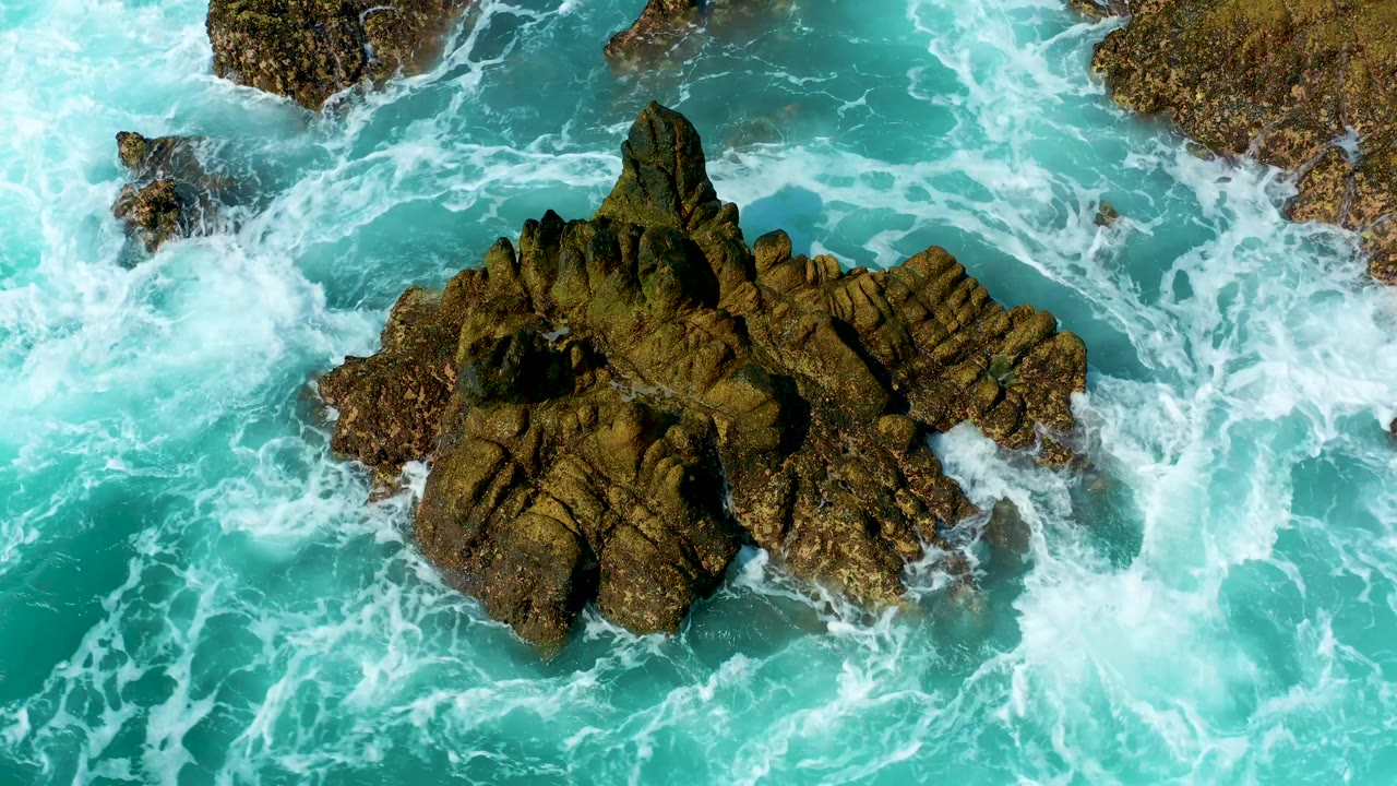 Aerial view in Marquis Los Cabos, Mexico, where thick white foam gathers along the lower edge of the jagged rock formation in bright turquoise water, with no visible people.