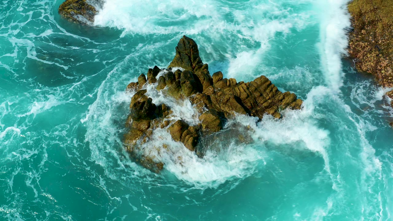 Overhead turquoise ocean waves breaking around a jagged rock outcrop.