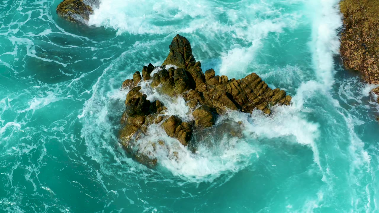 At Marquis Los Cabos, Mexico, white wave foam thickens on the upper and right sides of the centered rock outcrop, with no visible people.