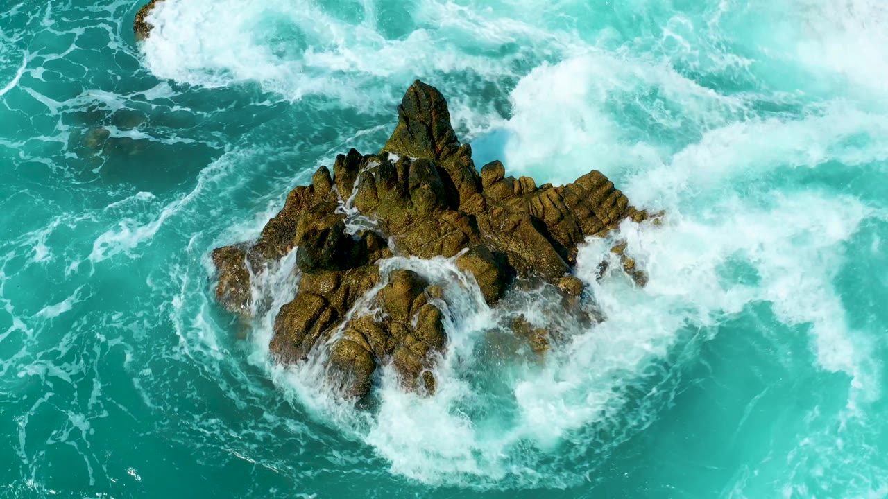 At Marquis Los Cabos, Mexico, the rock formation remains centered while a broad band of white surf spreads across the top of the turquoise water, with no visible people.
