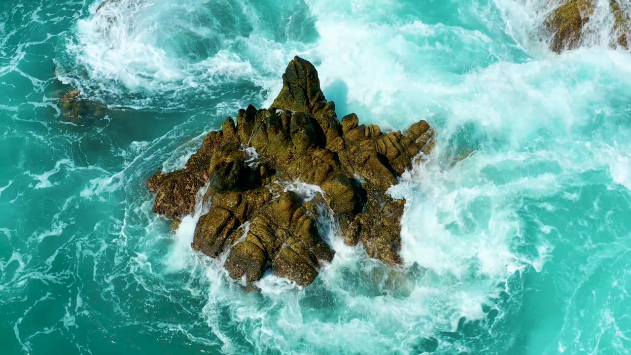 At Marquis Los Cabos, Mexico, bright foam surges along the right side of the central rock while calmer turquoise water remains at the lower right, with no visible people.