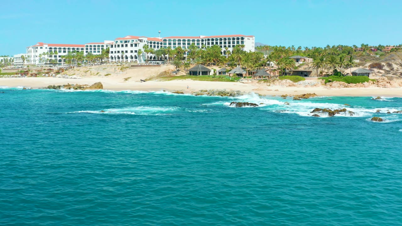 At 0:01, the Marquis Los Cabos shoreline remains centered with blue-green water in front, surf spreading around offshore rocks, palms on the beach, and no visible people.
