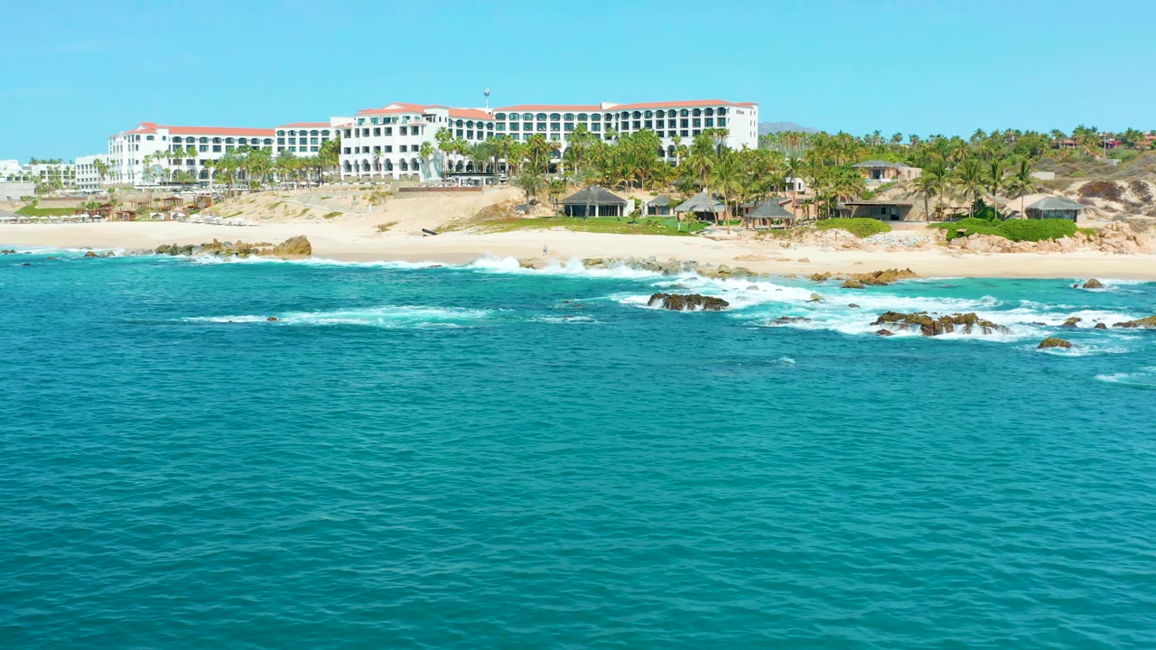 At 0:02, turquoise water fills most of the frame while white surf breaks near the sand below the Marquis Los Cabos building and palm-lined shore, with no visible people.