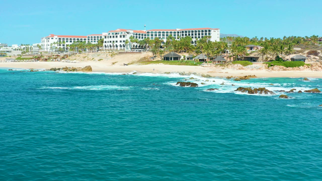 At 0:03, a wider stretch of the Marquis Los Cabos beach is visible, with surf breaking over rocks in the midground, white buildings behind the sand, and no visible people.