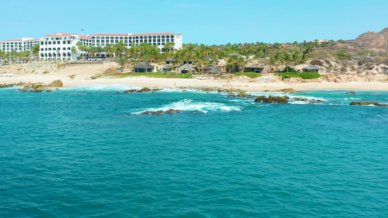One second in, the Marquis Los Cabos coastline remains wide in frame, with blue-green water in front, scattered dark rocks, a sandy beach, palms, and the white resort shifted near the upper left, no visible people.