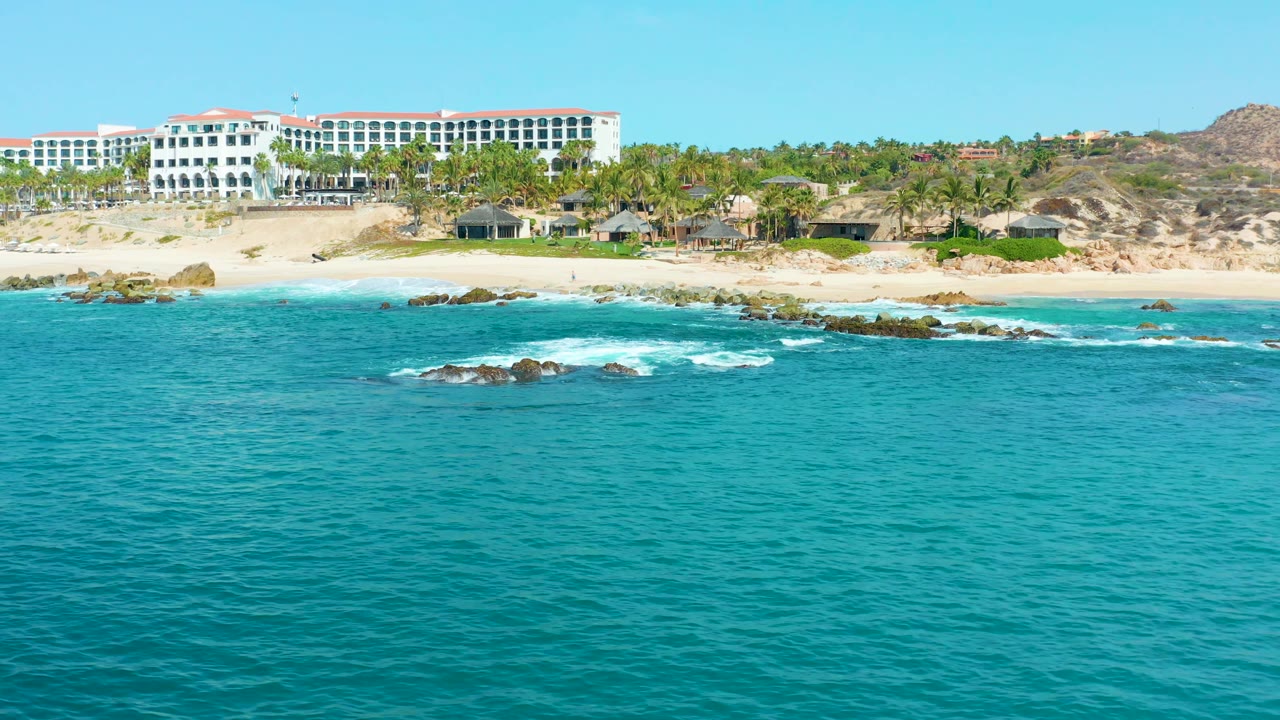 At 0:02, turquoise ocean and foamy water around low rocks lead to the sandy Marquis Los Cabos beach, palm trees, and a large white resort with red roof sections, no visible people.