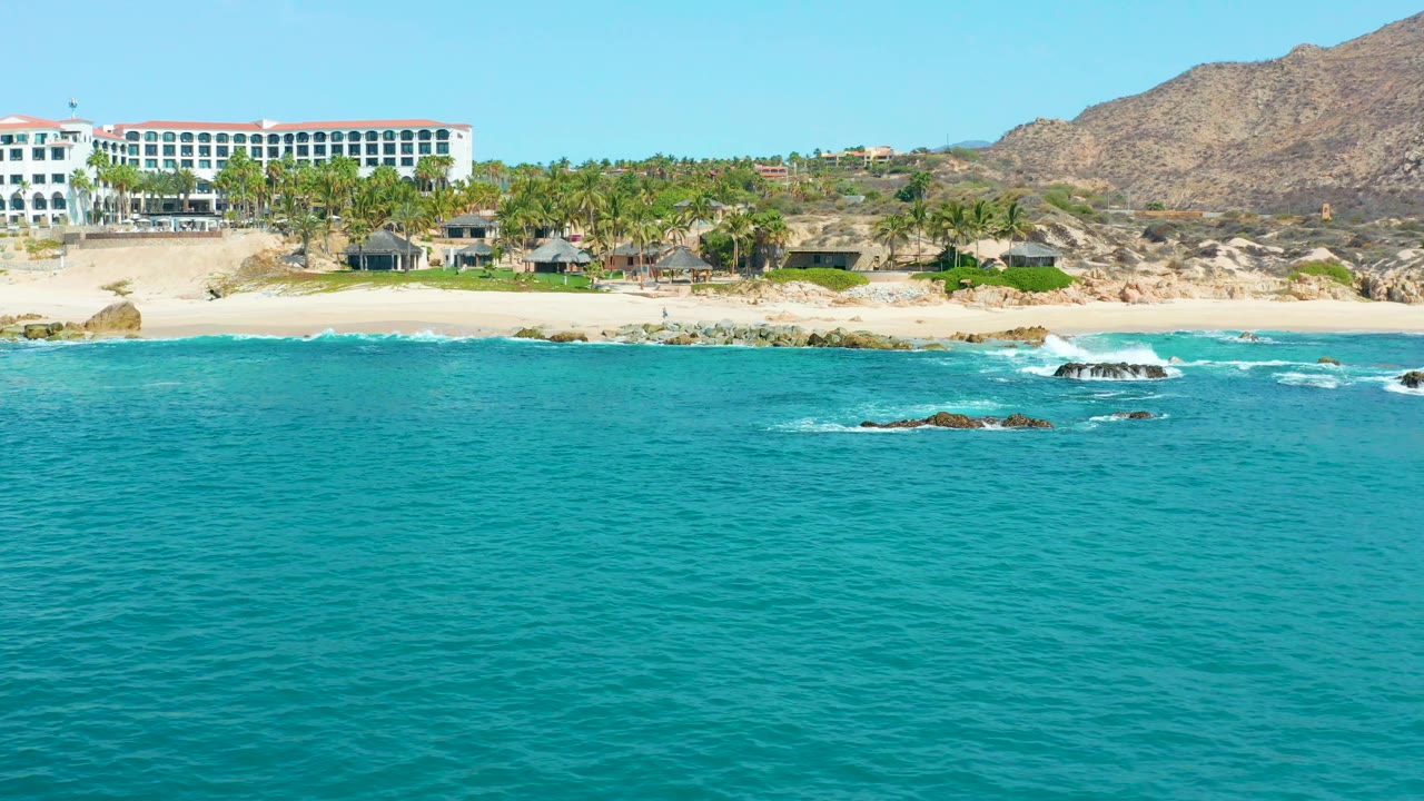 Turquoise ocean facing a sandy beach, rocks, palm trees, and a white resort.