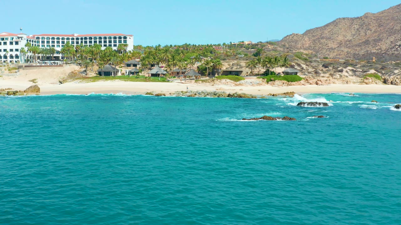 At 0:01, the Marquis Los Cabos shoreline stays wide while white foam gathers around offshore rocks, with the resort and palms running behind the sandy beach and no visible people.