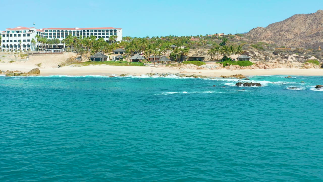 At 0:03, the sandy beach and white resort at Marquis Los Cabos sit above a rocky surf line, with low white waves crossing the turquoise water and no visible people.