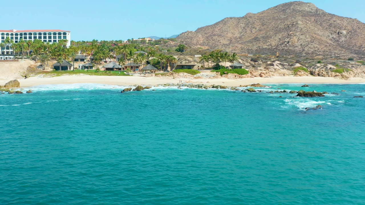 Wide elevated view at Marquis Los Cabos, Mexico with turquoise ocean in front, white surf on rocks near the beach, palm trees, coastal buildings, a white resort building, mountain backdrop, and no visible people.